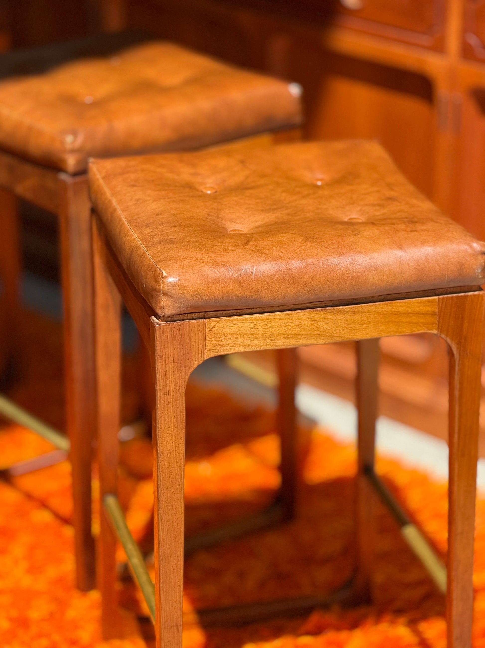 A set of four Gerald Easden 'Module' 1960s Blackwood bar stools stands on an orange shag carpet, with wood-paneled furniture in the background.