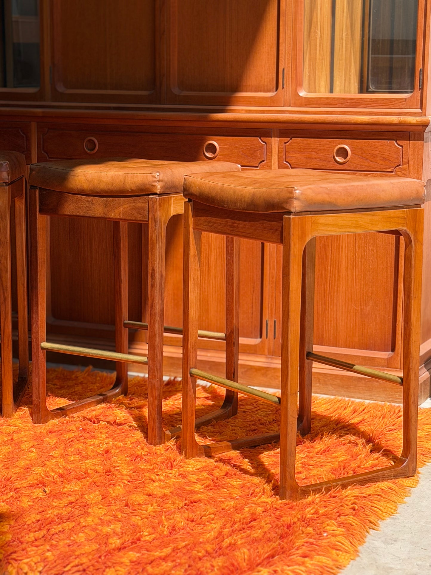 Four Gerald Easden ‘Module’ 1960s Blackwood Bar Stools (Revive) are arranged on an orange shag rug in front of a wooden cabinet, with sunlight casting elegant shadows across the scene.