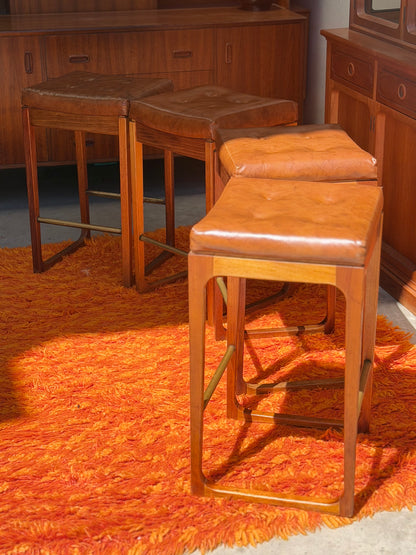 Four vintage Gerald Easden ‘Module’ 1960s Blackwood bar stools from Revive, with brown leather seats, stand on a bright orange shag rug amid mid-century modern wooden furniture, sunlight illuminating the inviting retro scene.