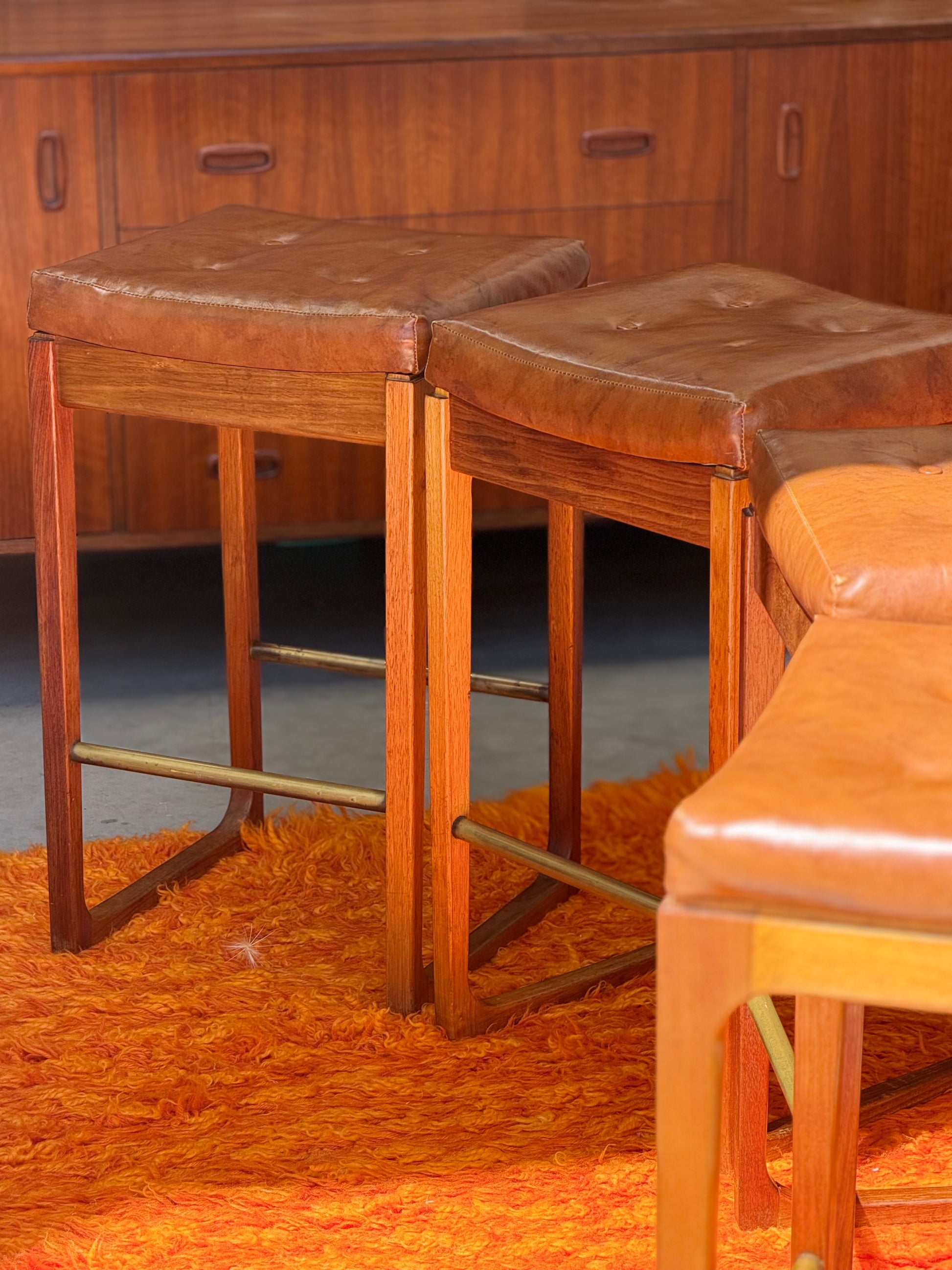 Four Gerald Easden ‘Module’ 1960s blackwood bar stools (Revive) sit on an orange shag rug, with a wooden sideboard in the background.