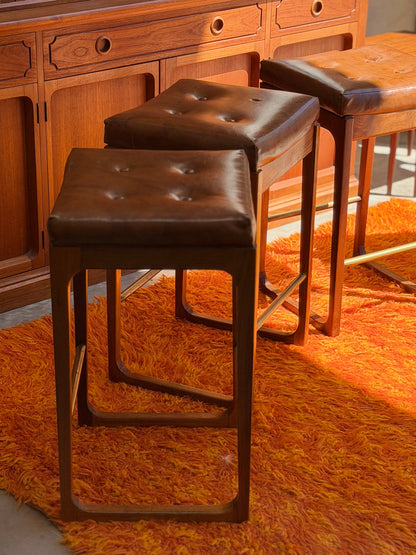 Four Gerald Easden ‘Module’ 1960s Blackwood Bar Stools (Revive) with brown tufted leather seats sit on a bright orange shag rug, in front of a paneled wooden cabinet, filling the room with warm mid-century modern charm and sunlight.