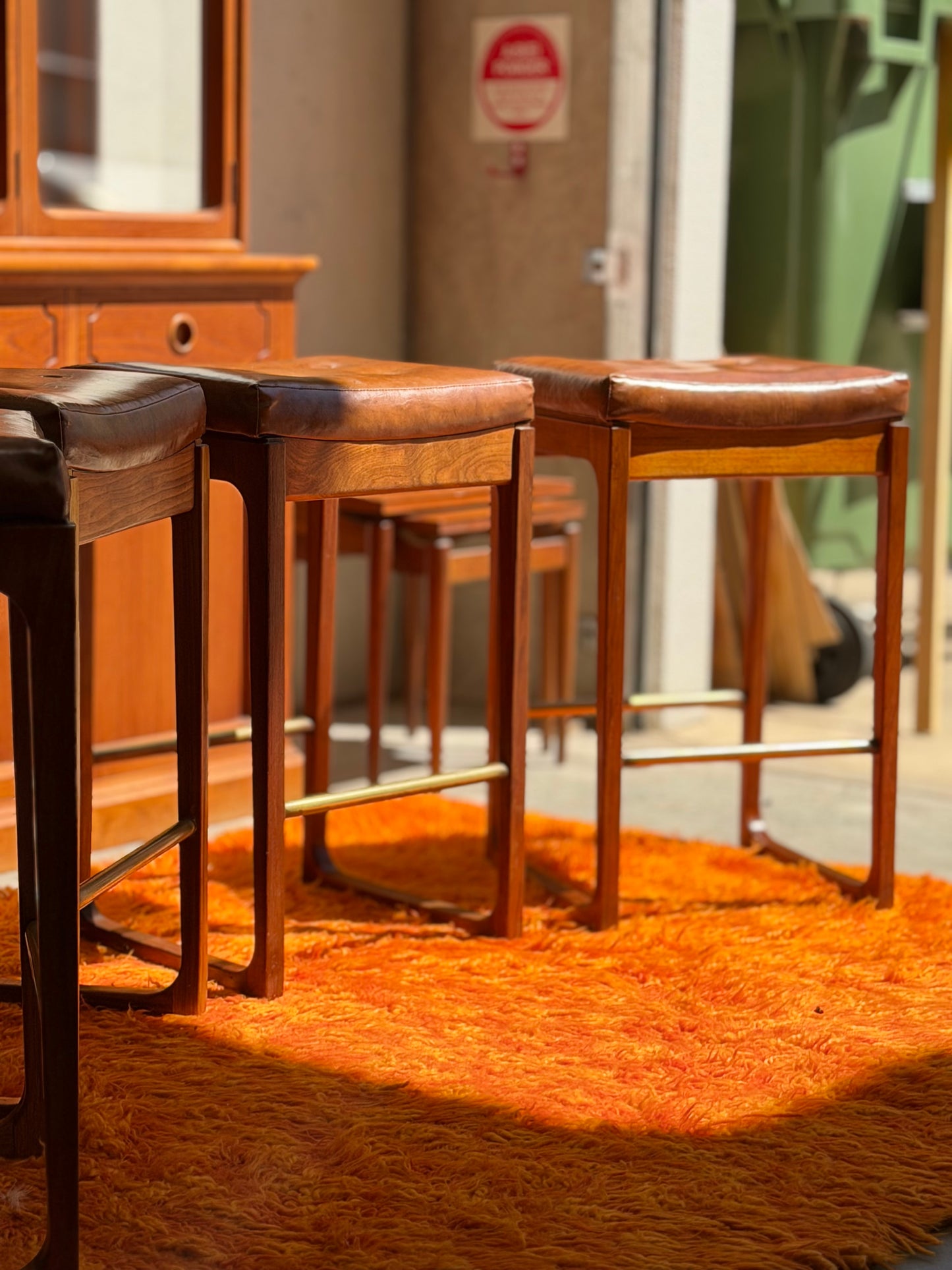 Four Gerald Easden ‘Module’ 1960s Blackwood bar stools from Revive sit on an orange shag rug in a sunlit room with wooden furniture and a broom visible in the background.
