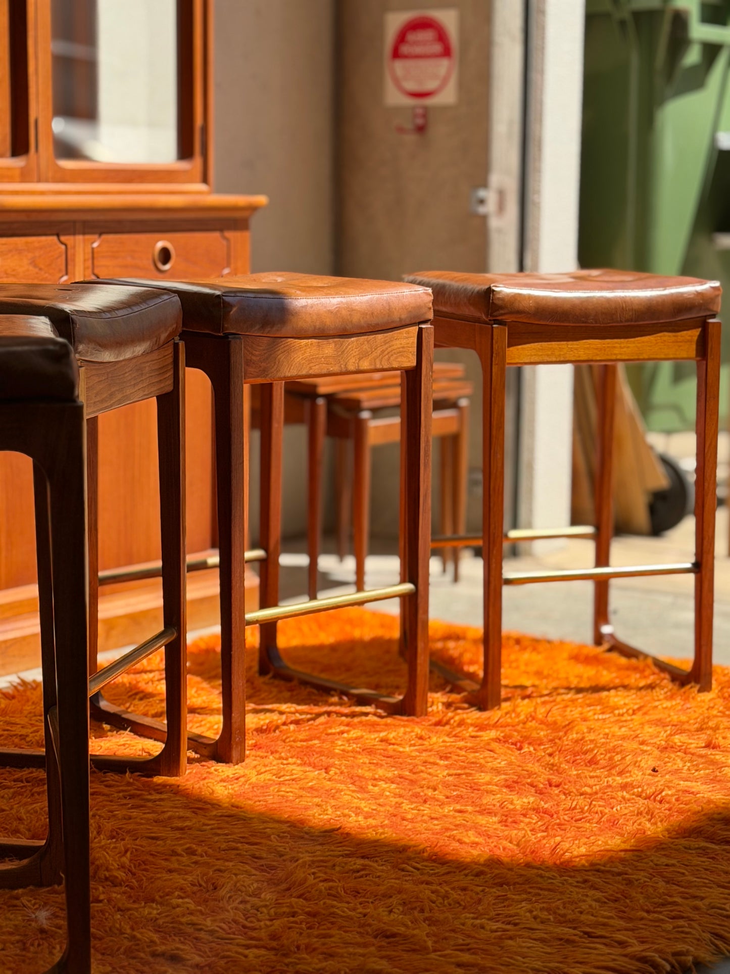 Four Gerald Easden 'Module' 1960s Blackwood Bar Stools sit on an orange shag rug in a sunlit room with wooden furniture and a No Smoking sign in the background.