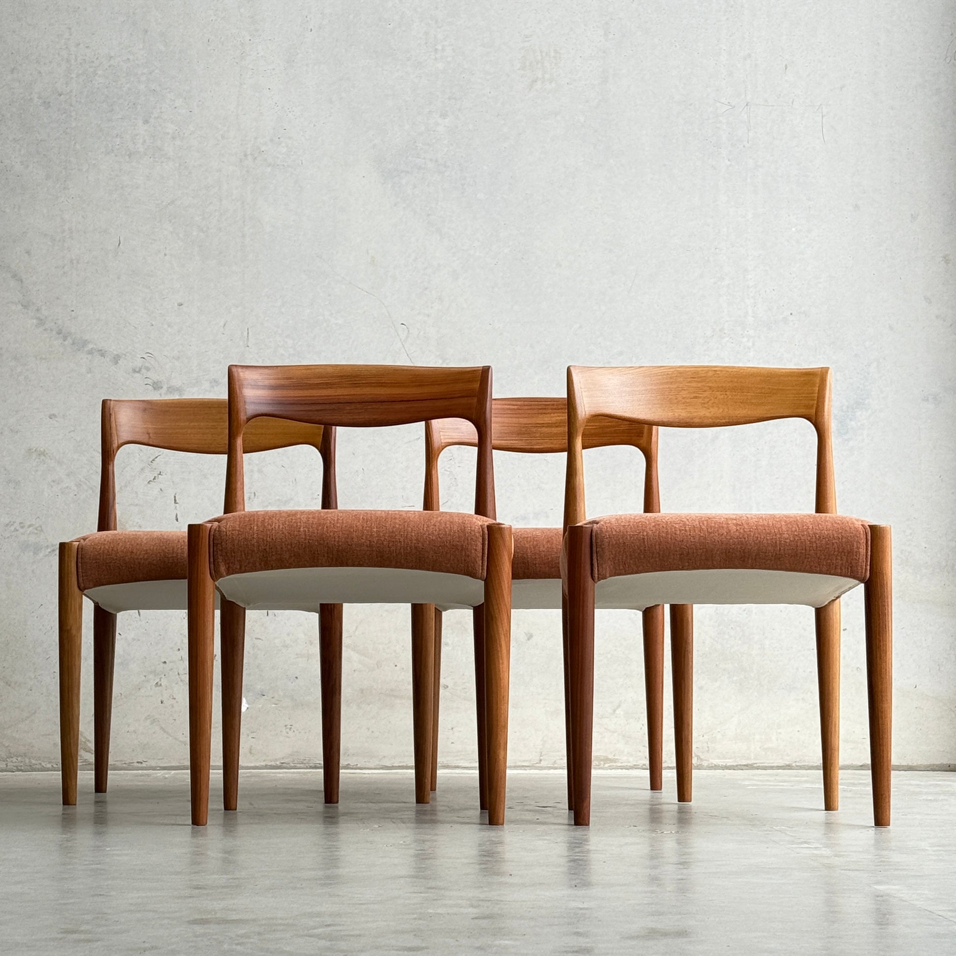 A set of four fully restored THB 'Milan' dining chairs (Rediscover), featuring minimalist mid-century modern design, is arranged in a staggered row against a plain light gray wall. Each chair has curved backrests and light brown upholstered seats.
