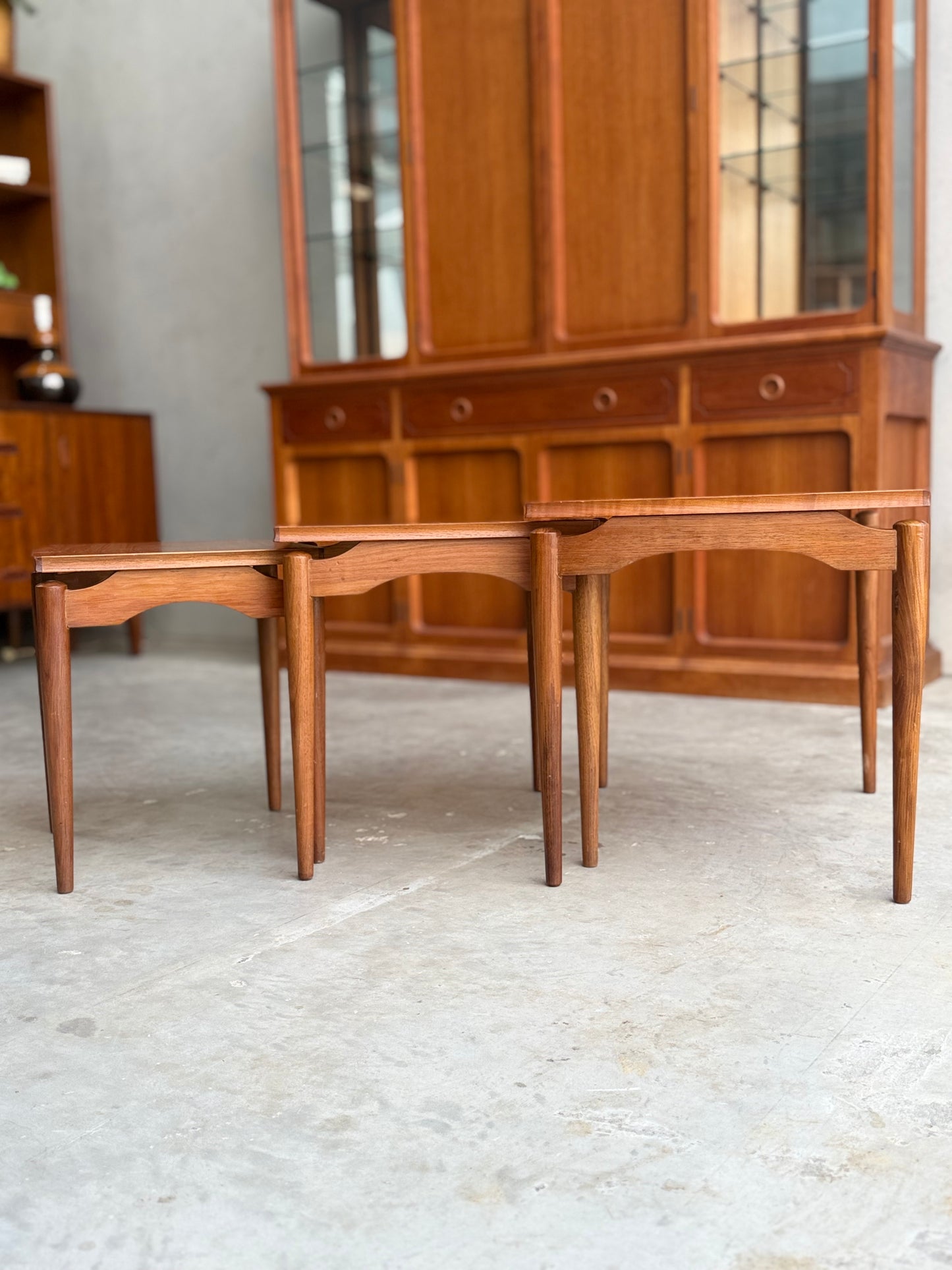 Two Noblett Mid-Century Floating Teak Nesting Tables – Circa 1960s (Revive) with tapered legs are placed side by side on a concrete floor, complemented by a hutch with glass doors and drawers for a Danish-inspired look.