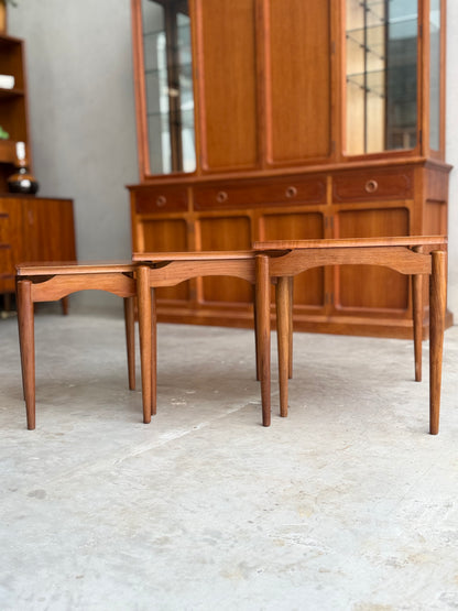 Two Noblett Mid-Century Floating Teak Nesting Tables – Circa 1960s (Revive) with tapered legs are placed side by side on a concrete floor, complemented by a hutch with glass doors and drawers for a Danish-inspired look.