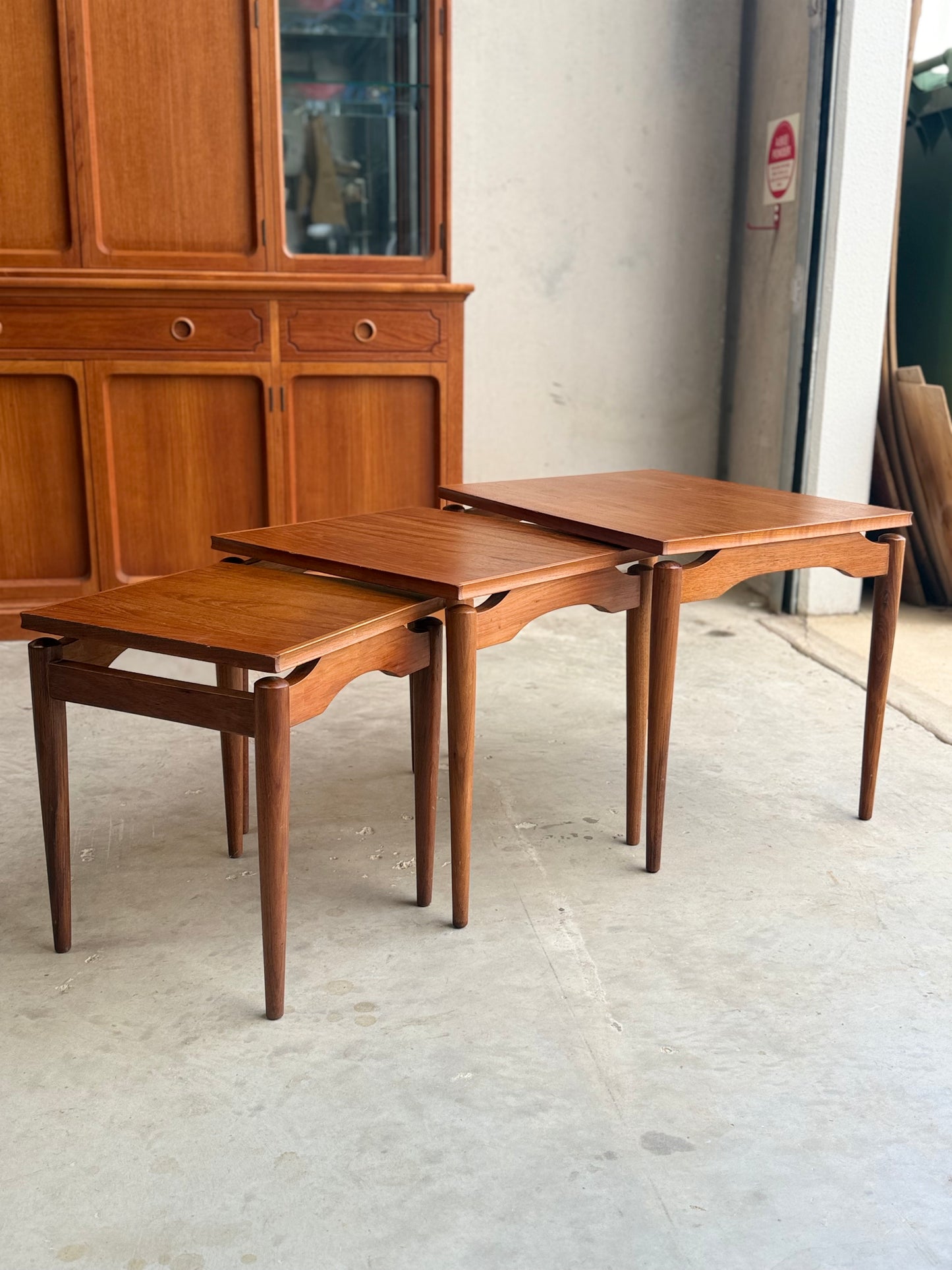 Three Noblett Mid-Century Floating Teak Nesting Tables – Circa 1960s (Revive) are stacked on a concrete floor, with a matching wooden cabinet in the background, highlighting classic Danish-inspired design.