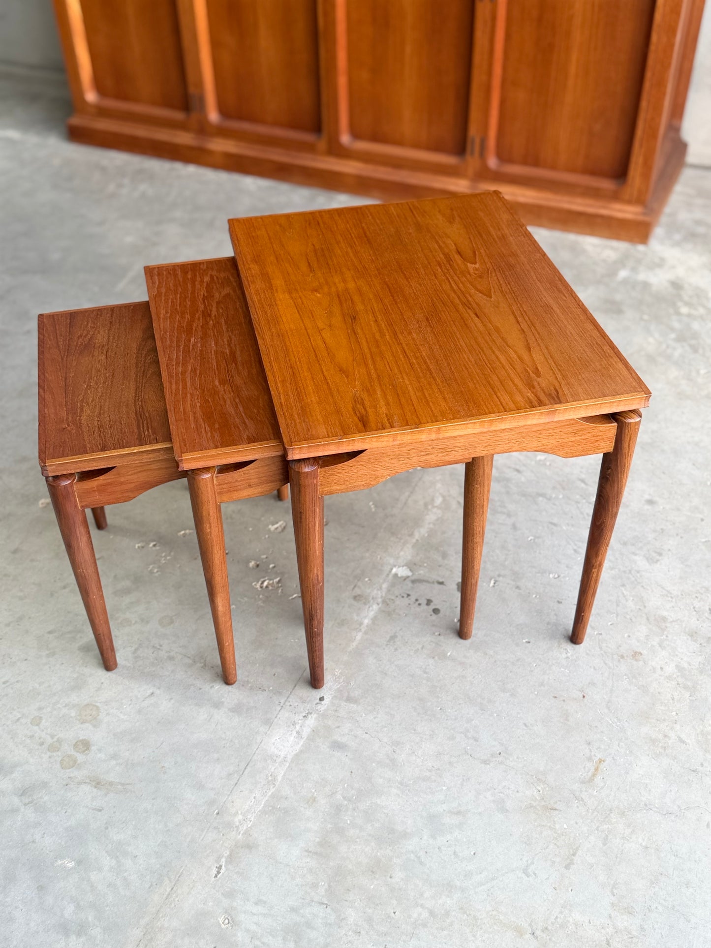 A set of Noblett Mid-Century Floating Teak Nesting Tables – Circa 1960s (Revive) is stacked on a concrete floor, showcasing their Danish-inspired design, with a wooden cabinet in the background.