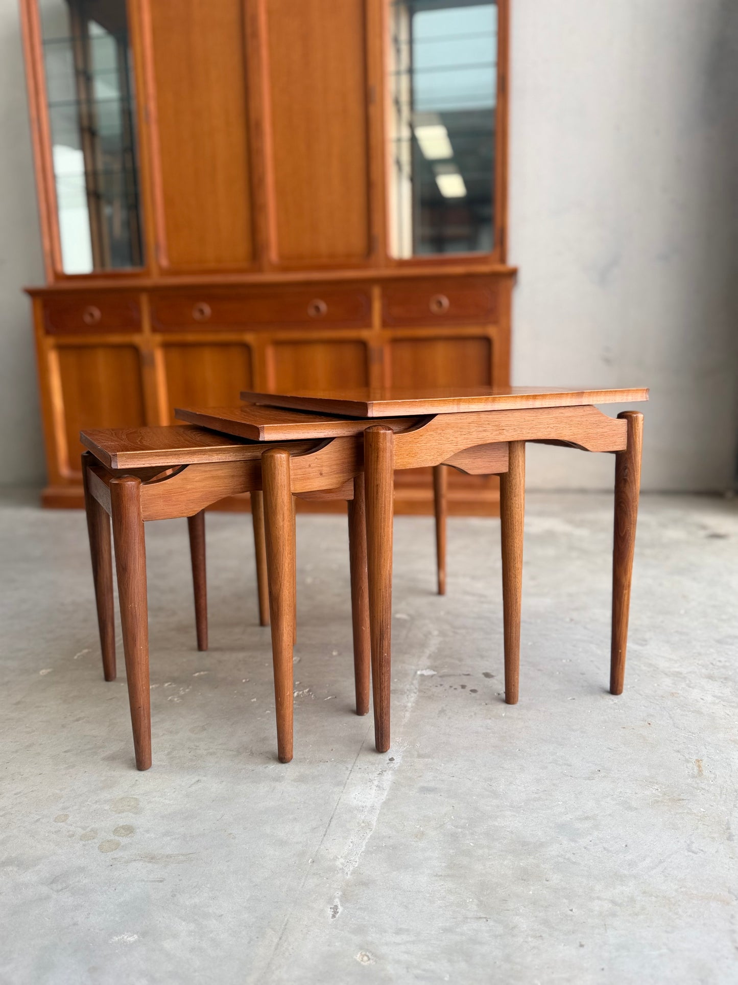 Three Noblett Mid-Century Floating Teak Nesting Tables – Circa 1960s (Revive) with tapered legs are arranged in front of a large cabinet with glass doors. The indoor scene features concrete flooring and neutral walls.