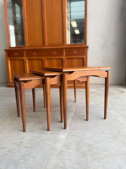 Three Noblett Mid-Century Floating Teak Nesting Tables – Circa 1960s (Revive) with tapered legs are arranged in front of a large cabinet with glass doors. The indoor scene features concrete flooring and neutral walls.
