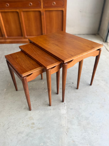 Three Noblett Mid-Century Floating Teak Nesting Tables – Circa 1960s (Revive) with Danish-inspired design and tapered legs are grouped on a concrete floor, accompanied by a matching wooden cabinet in the background.