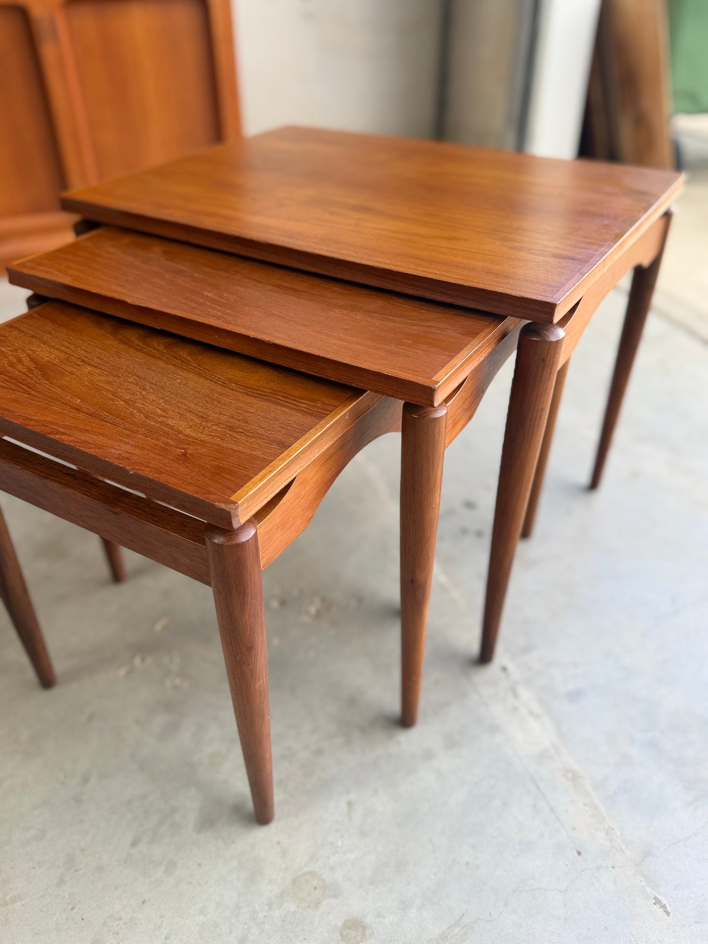 Three Noblett Mid-Century Floating Teak Nesting Tables – Circa 1960s (Revive) are stacked on a concrete floor in a well-lit room, showcasing their polished teak finish and classic Danish-inspired design.