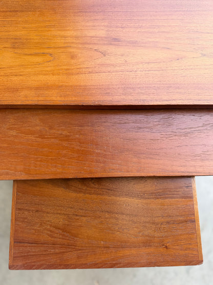 Close-up of three overlapping surfaces of the Noblett Mid-Century Floating Teak Nesting Tables – Circa 1960s (Revive), highlighting their warm brown finish, visible teak grain, and Danish-inspired mid-century design. The background is blurred.