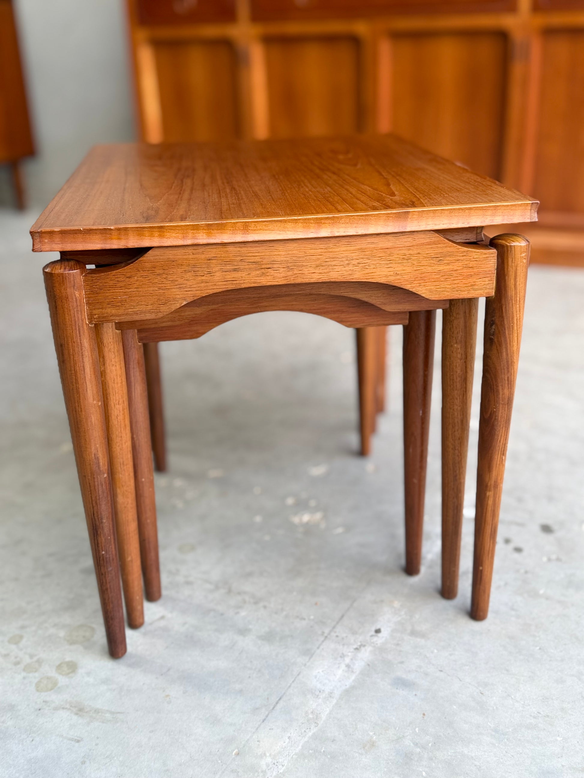 A set of Noblett Mid-Century Floating Teak Nesting Tables – Circa 1960s (Revive), with rounded legs and a smooth finish, radiates mid-century charm. Shown on a light concrete floor with wooden cabinets in the background.