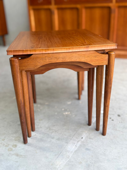 A set of Noblett Mid-Century Floating Teak Nesting Tables – Circa 1960s (Revive), with rounded legs and a smooth finish, radiates mid-century charm. Shown on a light concrete floor with wooden cabinets in the background.