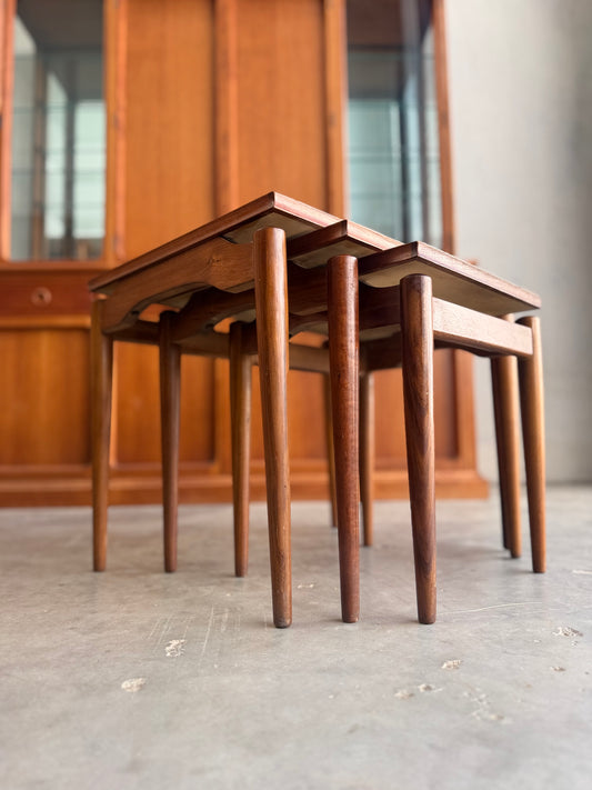 Three Noblett Mid-Century Floating Teak Nesting Tables – Circa 1960s (Revive) are stacked on a smooth concrete floor, set by a wooden cabinet. Their classic design brings timeless mid-century charm to any space.