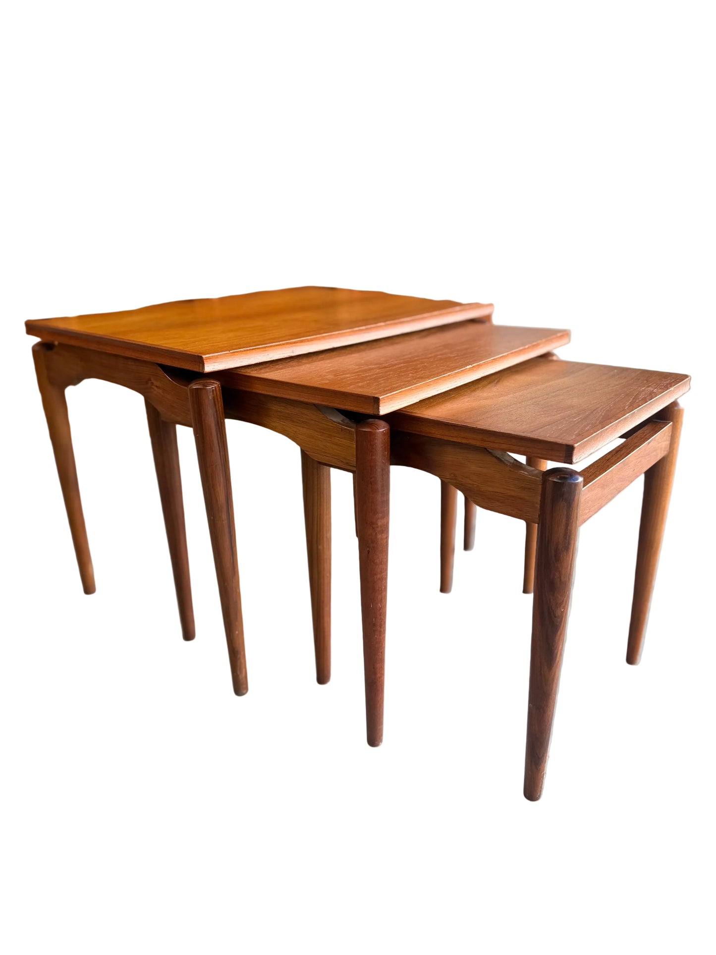 Three Noblett Mid-Century Floating Teak Nesting Tables – Circa 1960s (Revive) are stacked on a concrete floor, positioned in front of a matching wooden sideboard with drawers and glass-front cabinets.
