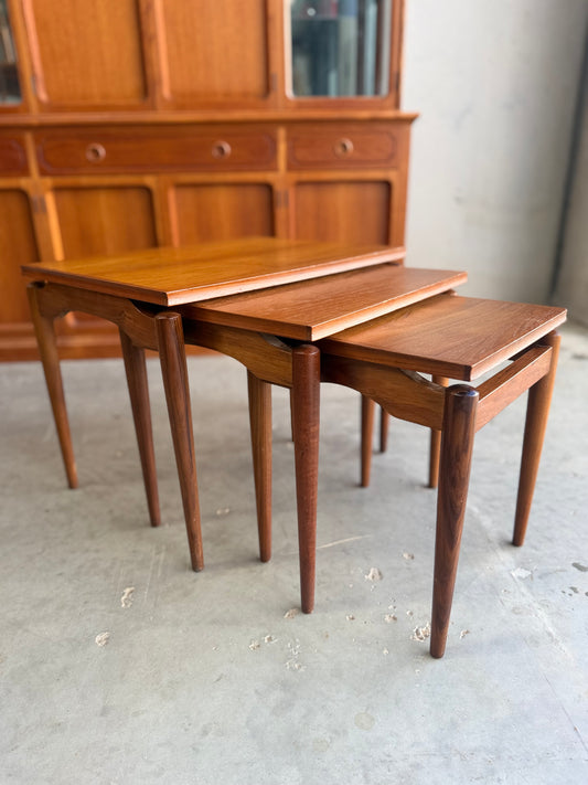 Three Noblett Mid-Century Floating Teak Nesting Tables – Circa 1960s (Revive) are stacked on a concrete floor, positioned in front of a matching wooden sideboard with drawers and glass-front cabinets.
