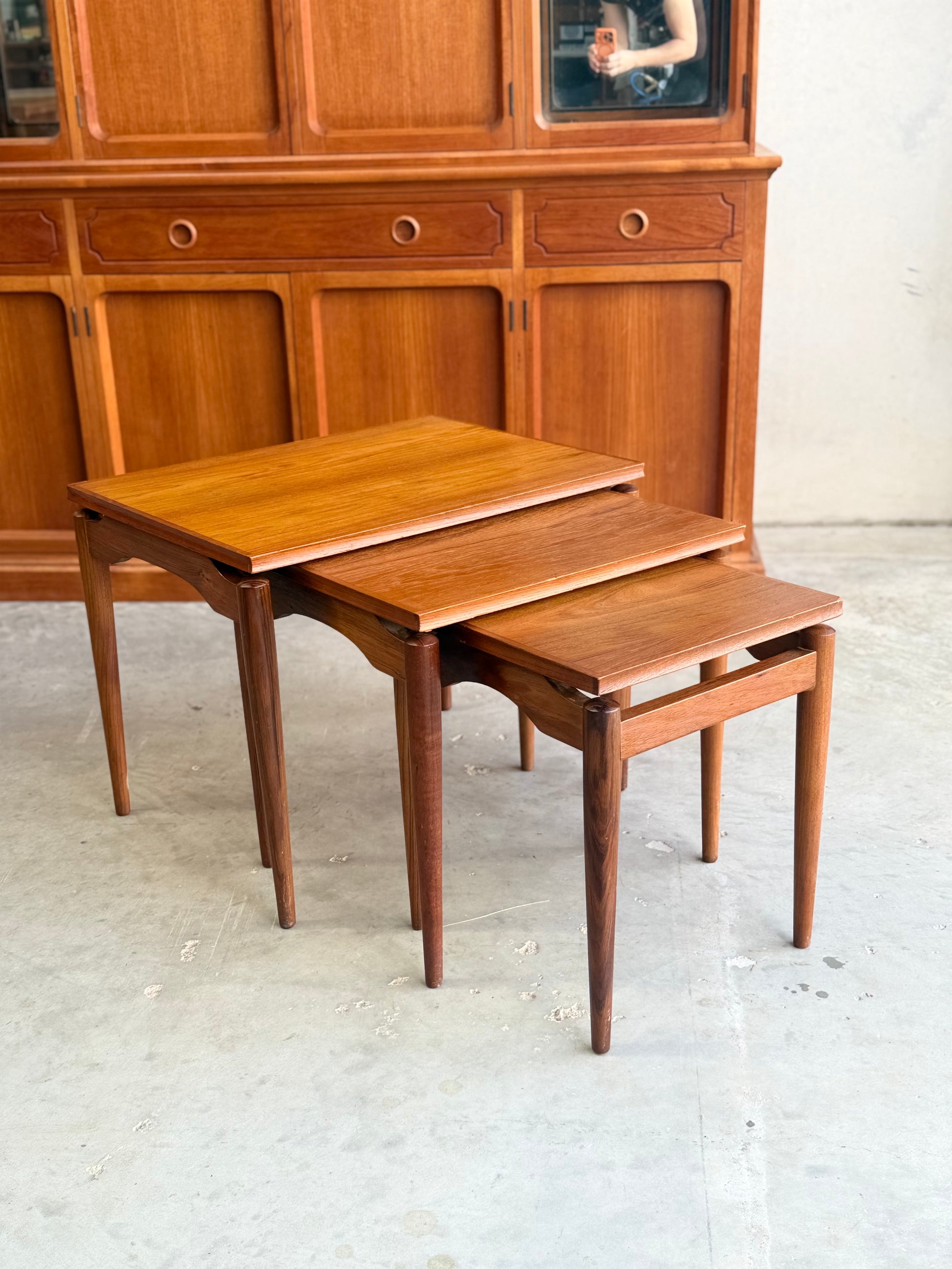 Three Noblett Mid-Century Floating Teak Nesting Tables – Circa 1960s (Revive) are stacked by a matching sideboard with drawers and cabinets, reflecting Danish-inspired style. A concrete floor and mirrored sideboard finish the scene.
