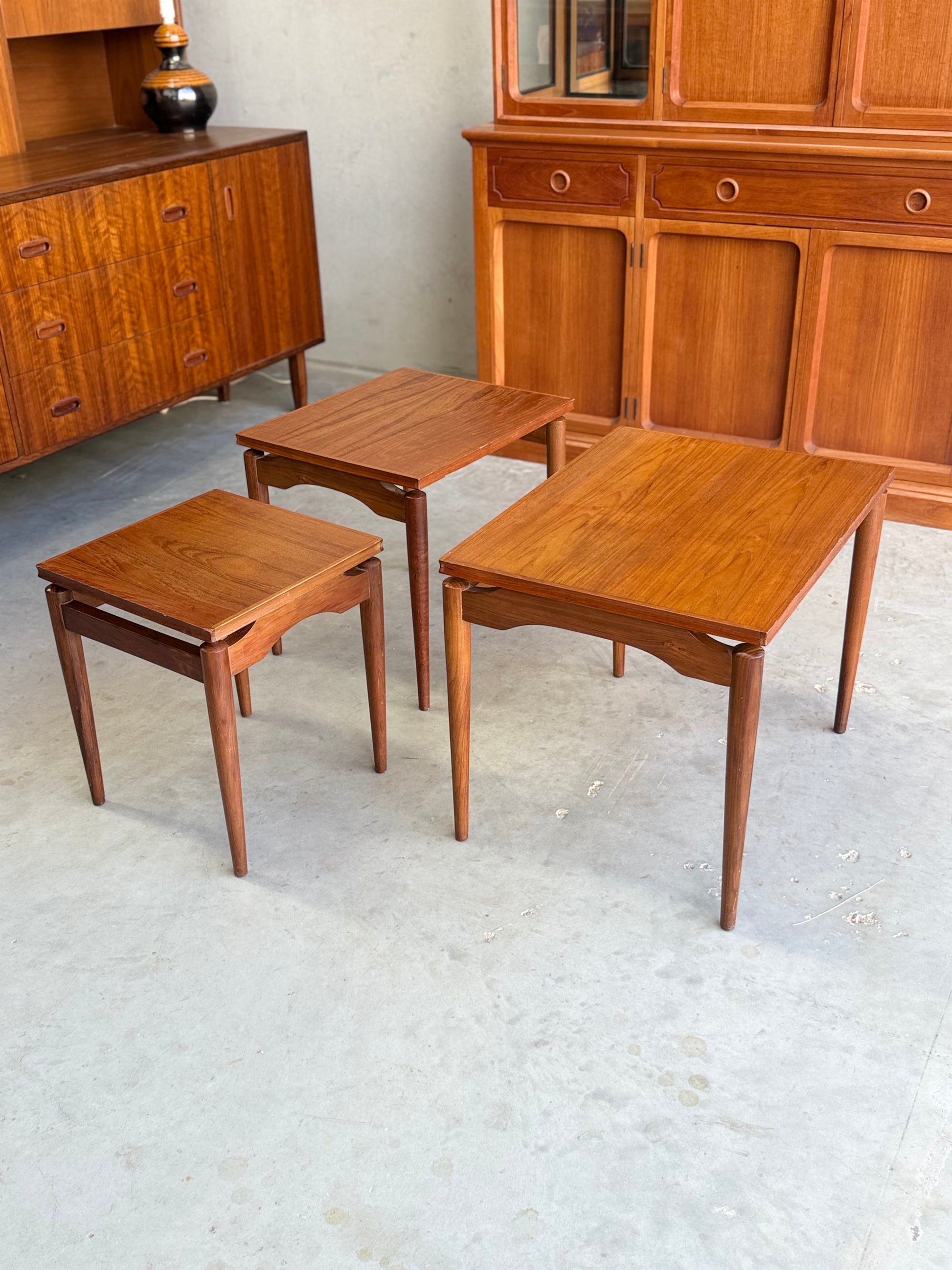 Three Noblett Mid-Century Floating Teak Nesting Tables – Circa 1960s (Revive) are arranged by size on a light concrete floor, with matching wooden cabinets and a lamp in the background, highlighting their Danish-inspired design.