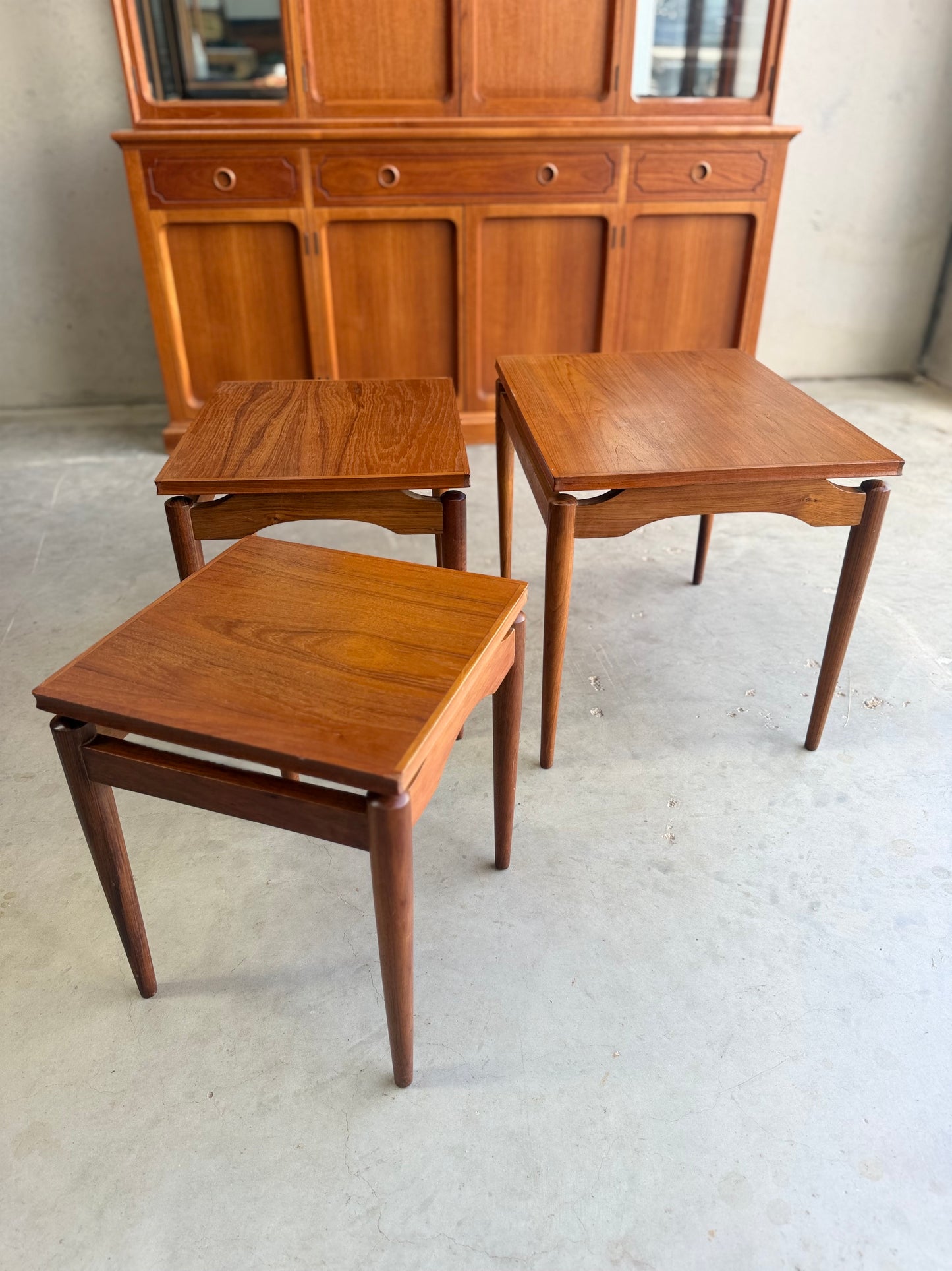 Three Noblett Mid-Century Floating Teak Nesting Tables – Circa 1960s (Revive) with tapered legs sit on a light concrete floor, complemented by a matching wooden sideboard featuring drawers and panel detailing in the background.