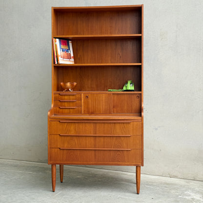 The Danish Teak Bureau Bookcase Secretary Desk by Erik Petersen for EP Møbler features open shelves, small drawers, and a pull-down desk, elegantly displaying books and décor against a concrete wall.