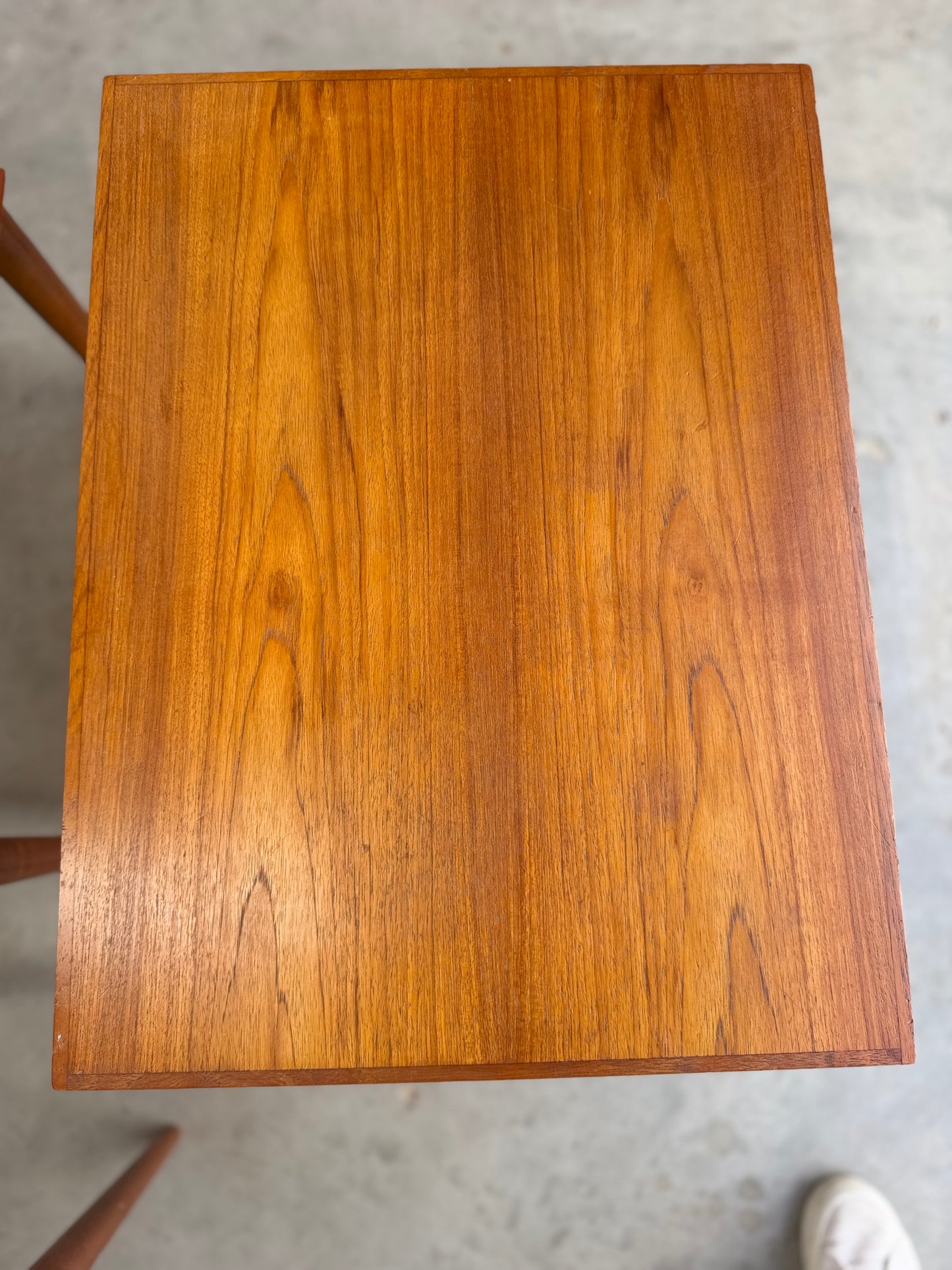 A rectangular teak table with a warm brown finish and visible grain stands on a light floor. The edge of another Noblett Mid-Century Floating Teak Nesting Table – Circa 1960s (Revive) and part of a white shoe appear in the corners.