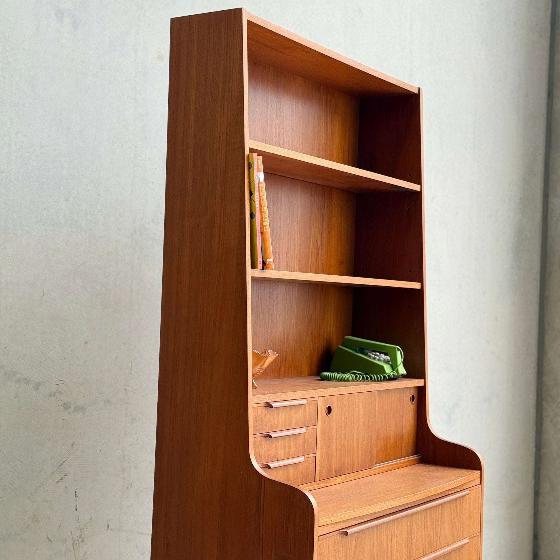 The Danish Teak Bureau Bookcase Secretary Desk by Erik Petersen for EP Møbler features open shelves, a built-in desk, and small drawers in Scandinavian style, with books and green organizers arranged against a plain light wall.