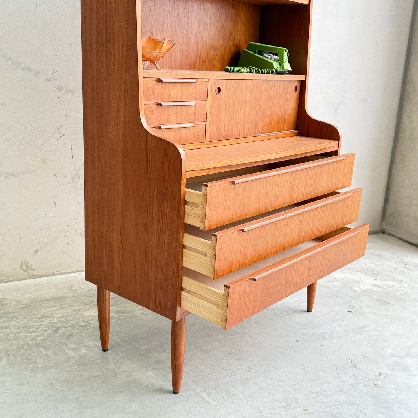 A Danish Teak Bureau Bookcase Secretary Desk by Erik Petersen for EP Møbler features tapered legs, three partially open drawers, and upper shelves with a ceramic bird and green books, set against a plain concrete wall.