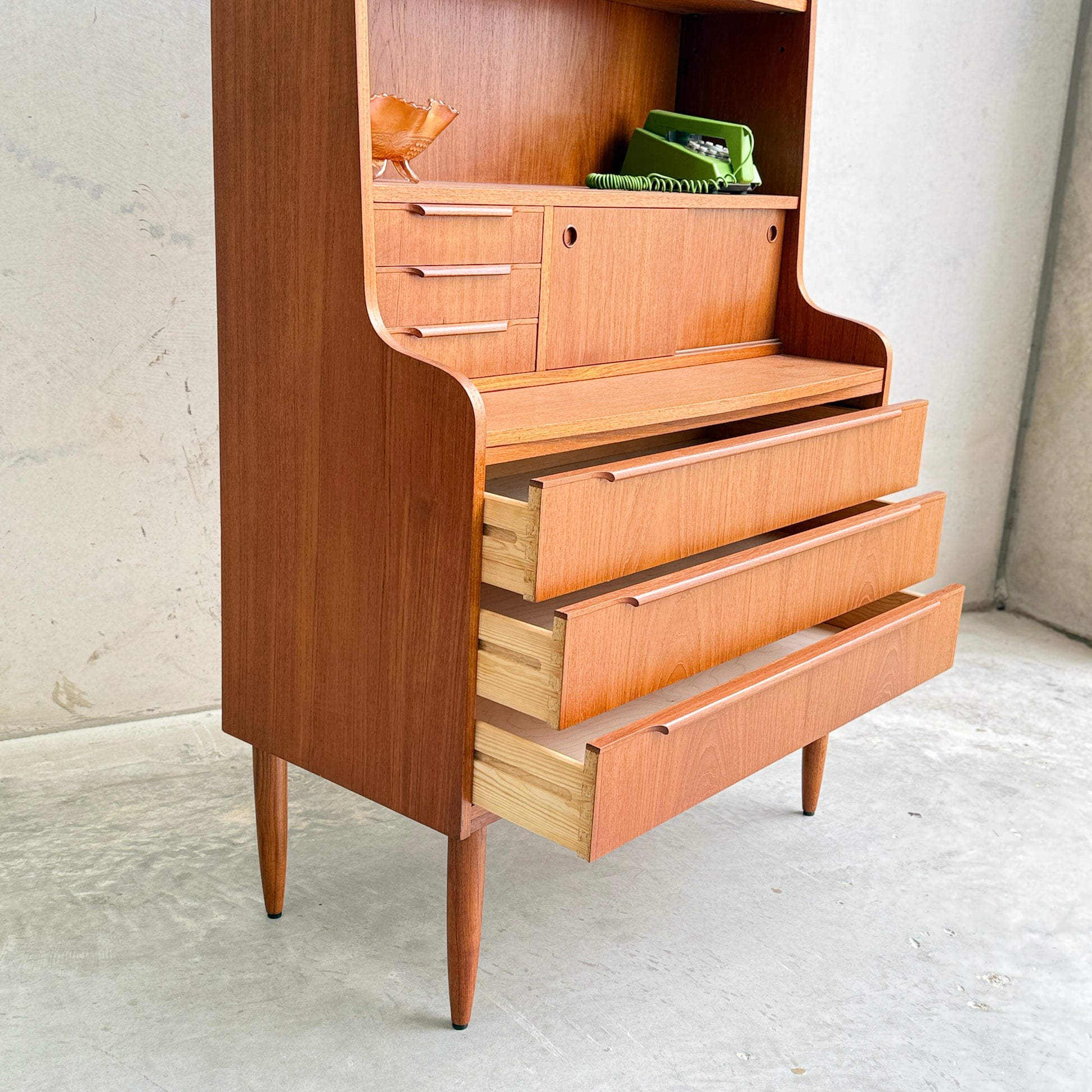 A Danish Teak Bureau Bookcase Secretary Desk by Erik Petersen for EP Møbler features tapered legs, three partially open drawers, and upper shelves with a ceramic bird and green books, set against a plain concrete wall.