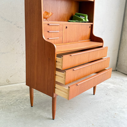 A Danish Teak Bureau Bookcase Secretary Desk by Erik Petersen for EP Møbler features tapered legs, three partially open drawers, and upper shelves with a ceramic bird and green books, set against a plain concrete wall.