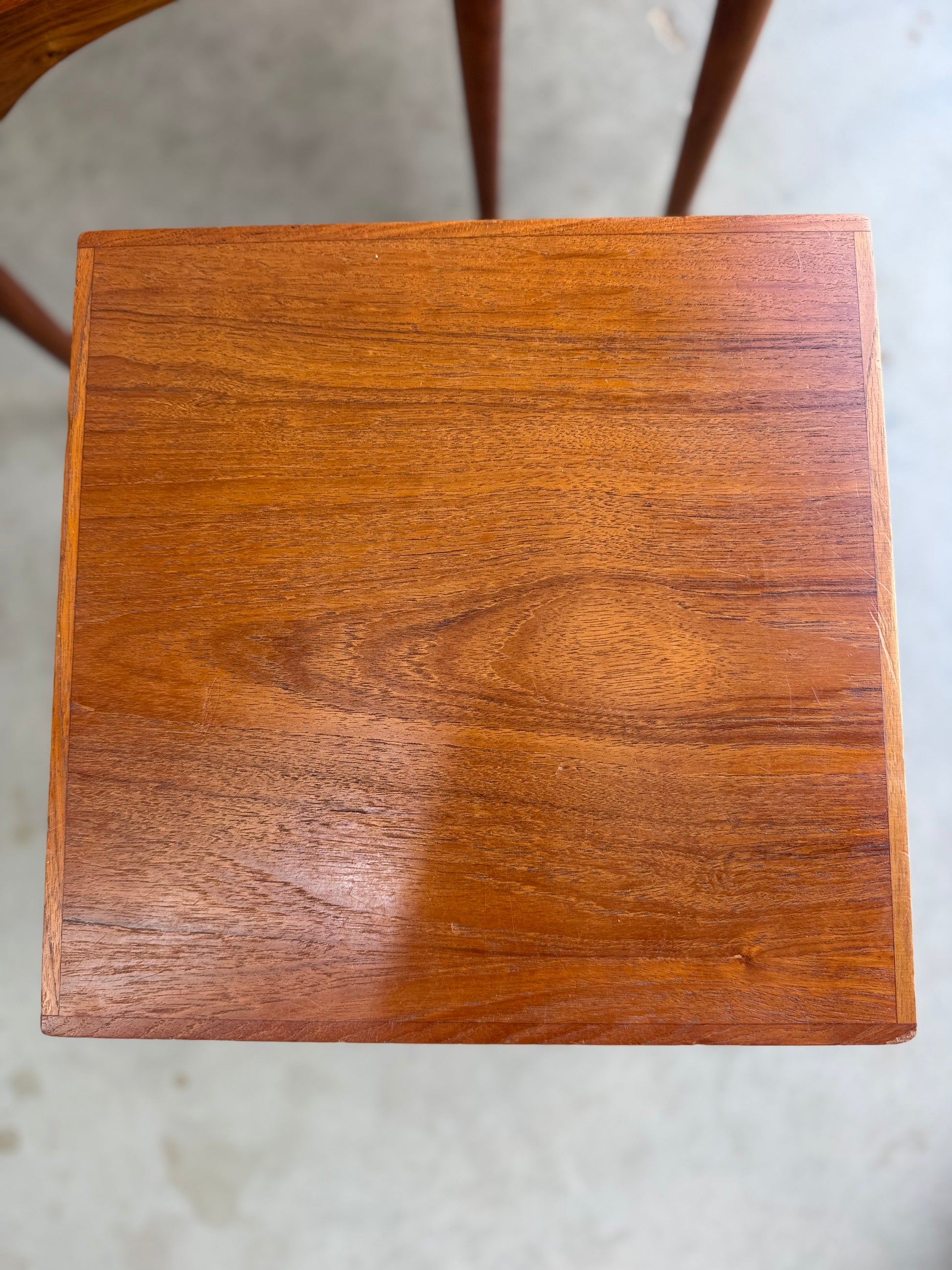 A close-up of the Noblett Mid-Century Floating Teak Nesting Table (Revive, circa 1960s) shows a square wooden top with visible grain, smooth polished surface, and natural brown finish, evoking classic Danish-inspired design on a concrete floor.