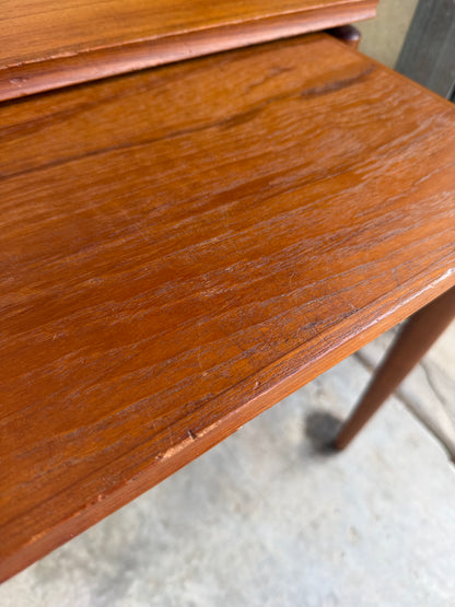 A close-up of a Noblett Mid-Century Floating Teak Nesting Table – Circa 1960s (Revive) shows its smooth reddish-brown finish with visible scratches, minor edge wear, and slender mid-century legs standing on a concrete floor.