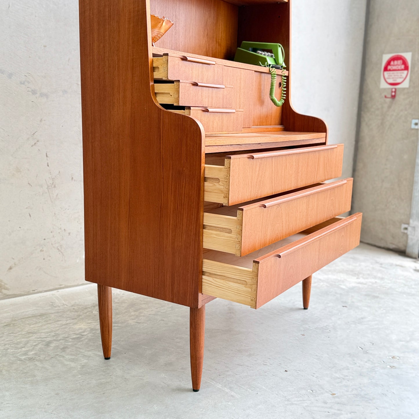 The Danish Teak Bureau Bookcase Secretary Desk by Erik Petersen for EP Møbler, with slim tapered legs and open drawers, stands on a concrete floor. A green rotary phone and book rest on top, evoking classic Scandinavian style. A fire alarm is seen in the background.