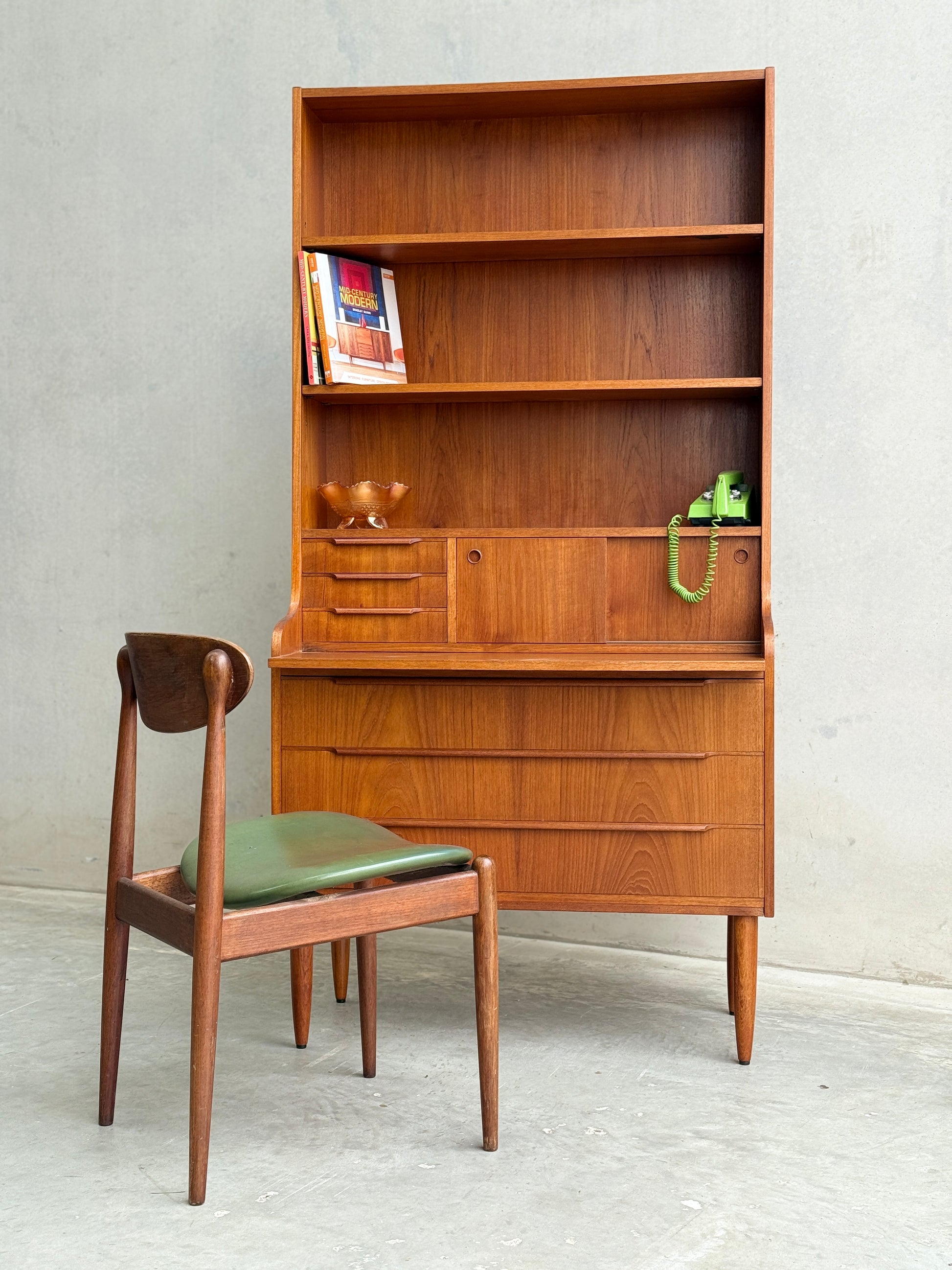 A Danish Teak Bureau Bookcase Secretary Desk by Erik Petersen for EP Møbler features shelves, drawers, an attached green rotary phone, and a wooden chair with a green seat in front; several books sit on the top shelf.