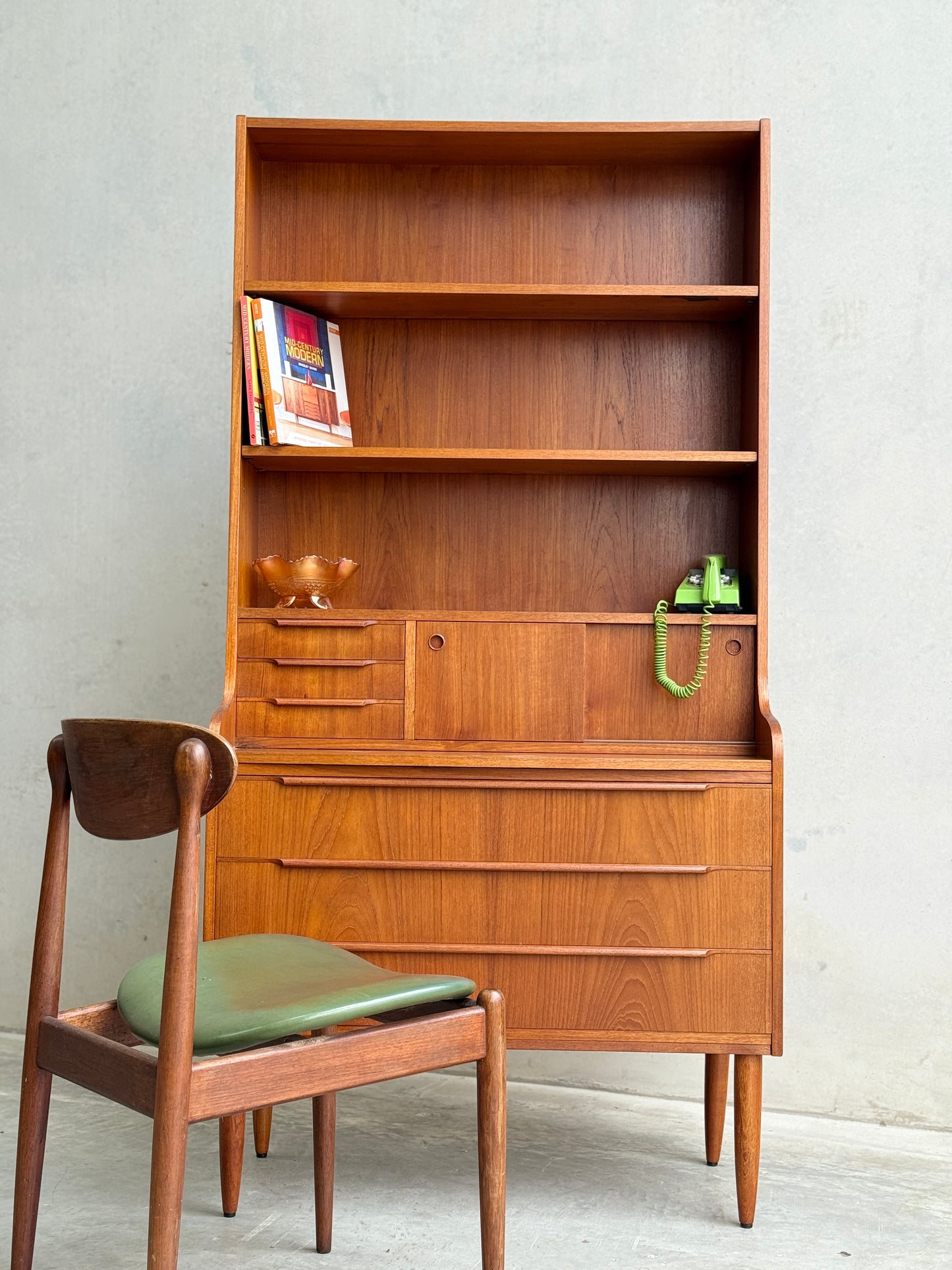 The Danish Teak Bureau Bookcase Secretary Desk by Erik Petersen for EP Møbler (Refine) features shelves, drawers, books, a green vintage telephone, glass bowl accents, and is paired with a matching wooden chair with a green cushion.