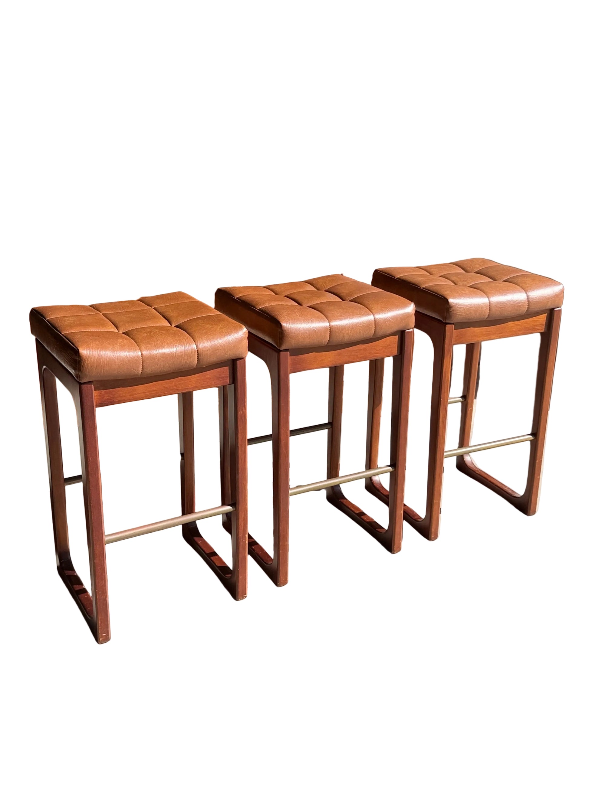 A set of three Mid Century Australian 1960s Barstools by Gerald Easden (Bar Height, Revive) with brown leather and wood, featuring square woven seats, are lined up on a sunlit concrete floor before a wooden glass-door cabinet.