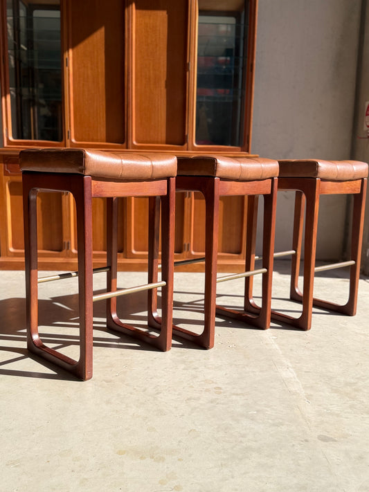 A set of three Mid Century Australian 1960s Barstools by Gerald Easden (Bar Height, Revive) features brown leather seats and wooden frames in a row on a sunlit floor, with wood cabinetry behind—a classic mid-century accent.