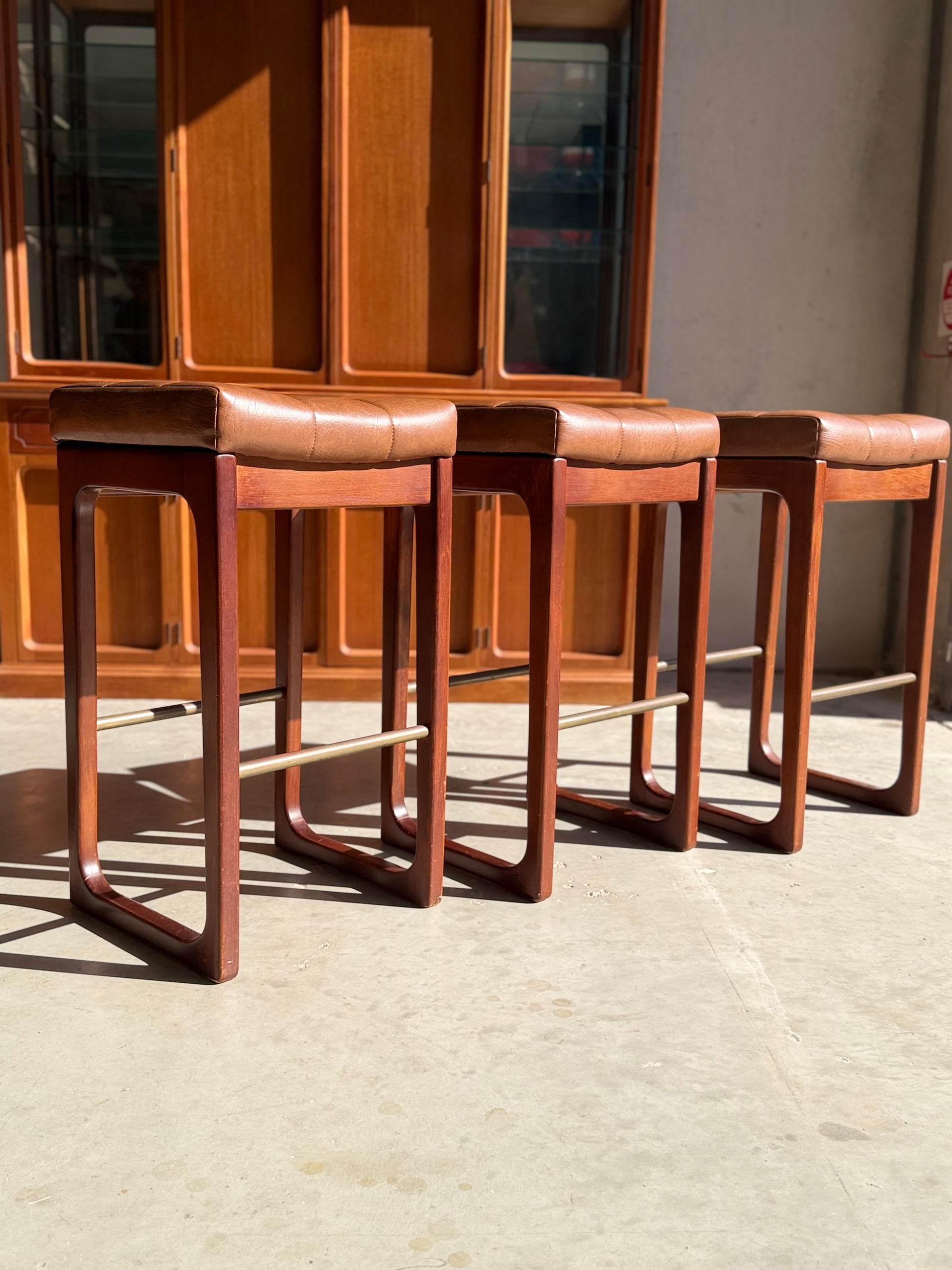 A set of three Mid Century Australian 1960s Barstools by Gerald Easden (Bar Height, Revive) features brown leather seats and wooden frames in a row on a sunlit floor, with wood cabinetry behind—a classic mid-century accent.