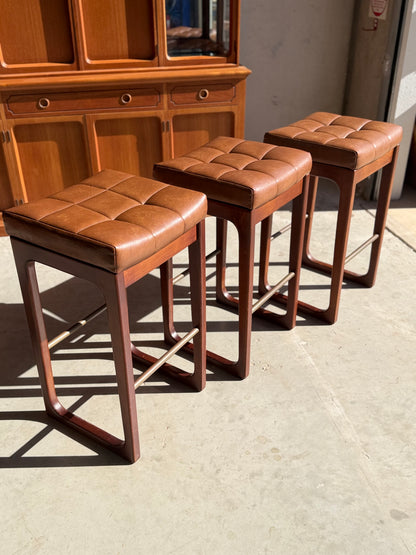 A set of three Mid Century Australian 1960s barstools by Gerald Easden (Bar Height, Revive) with brown tufted leather seats and wooden frames are lined up on a concrete floor, sunlight casting soft shadows, with a matching cabinet behind.
