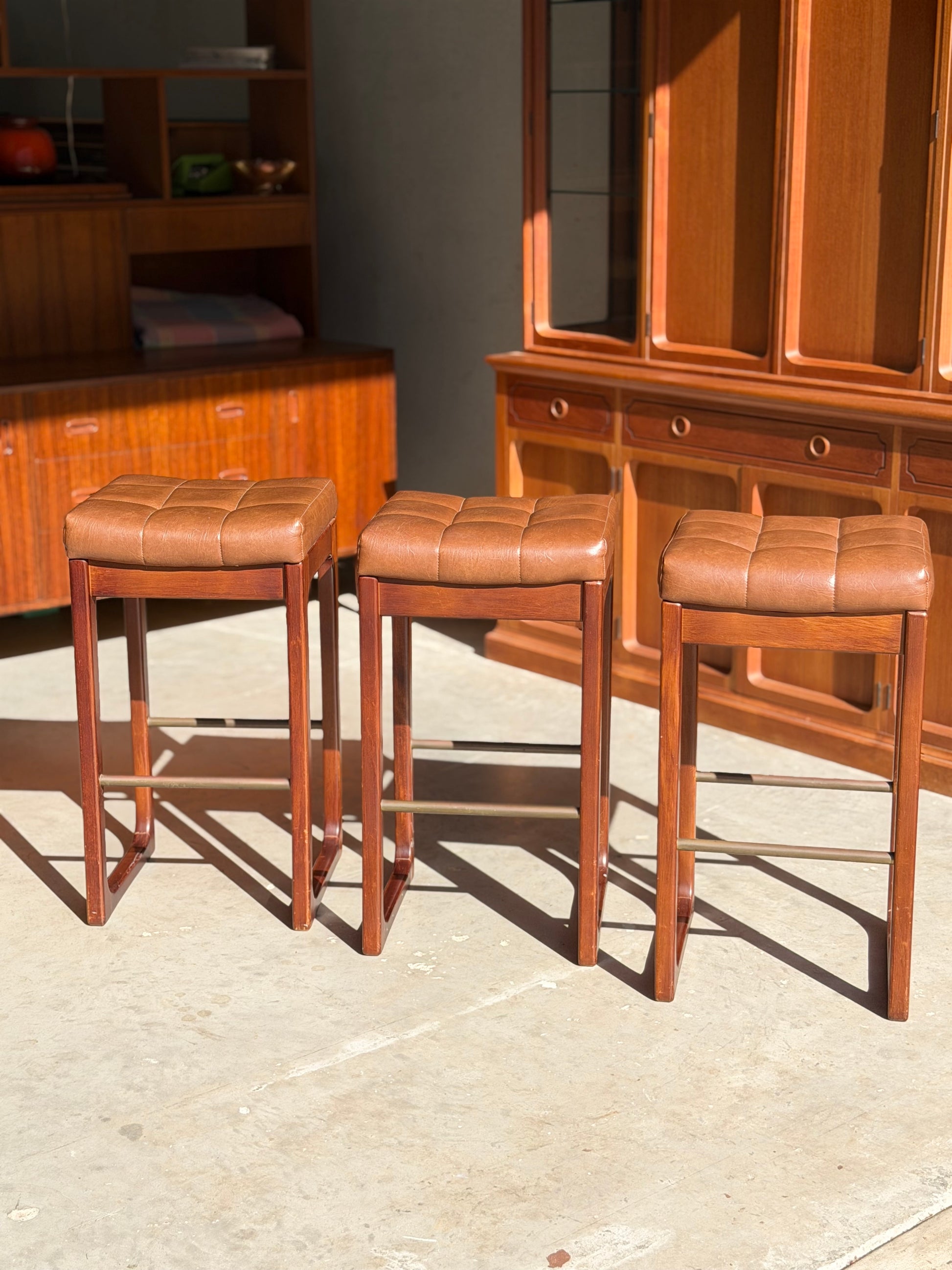 A set of three Mid Century Australian 1960s Barstools by Gerald Easden (Bar Height, Revive) are arranged in a row on a sunlit floor, with wooden cabinets and shelves in the background.