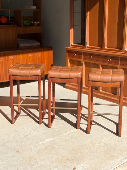 A set of three Mid Century Australian 1960s Barstools by Gerald Easden (Bar Height, Revive) are arranged in a row on a sunlit floor, with wooden cabinets and shelves in the background.