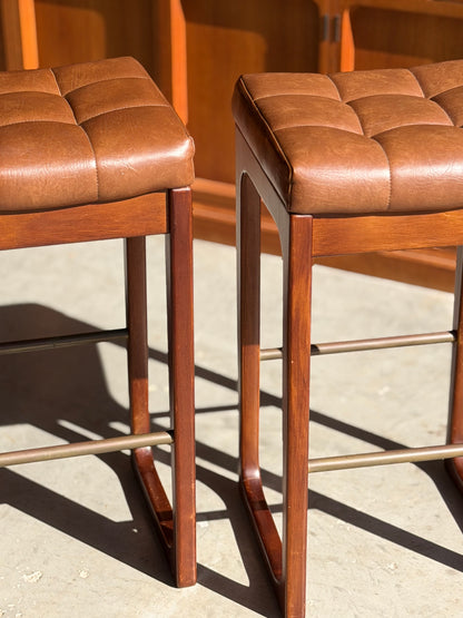 Three Gerald Easden Mid Century Australian 1960s barstools (Bar Height, Revive) with wooden frames and brown tufted leather seats sit side by side on a sunlit concrete floor, embodying timeless mid-century style.