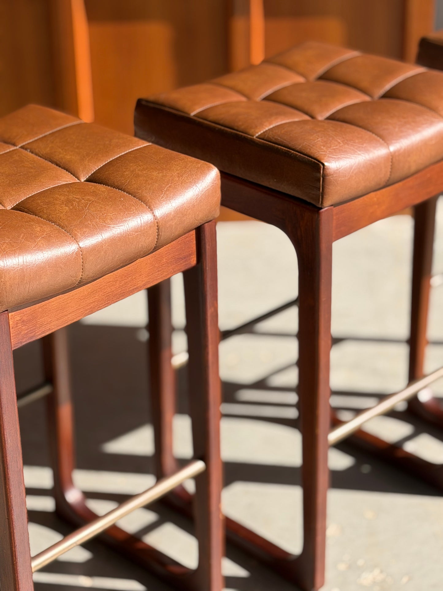 Set of 3 Mid Century Australian 1960s Barstools by Gerald Easden (Bar Height, Revive), featuring brown tufted leather seats and wooden legs, shown in sunlight casting distinct shadows on the floor.