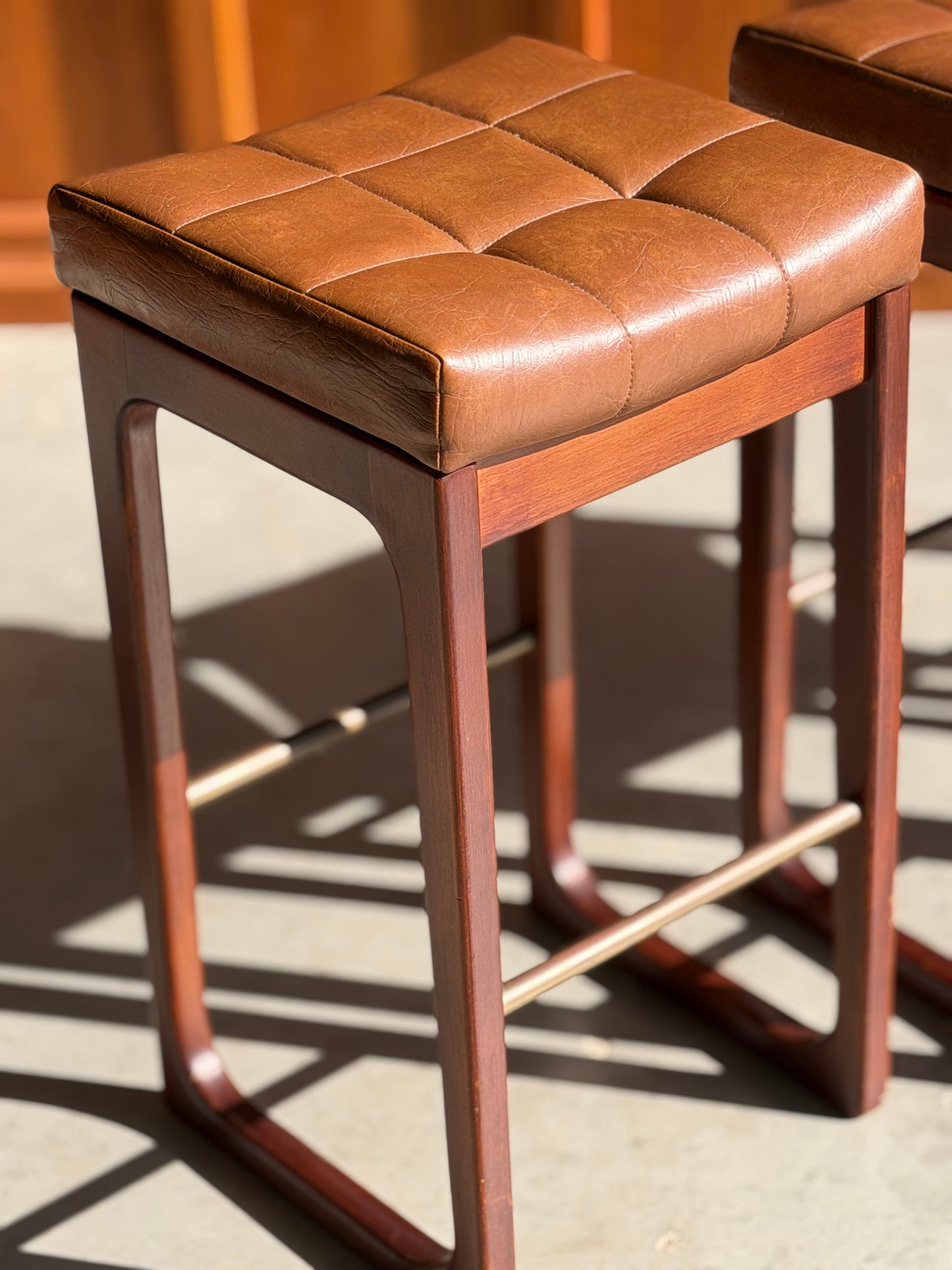 The Mid Century Australian 1960s Barstools by Gerald Easden (Set of 3 - Bar Height, Revive) feature square tufted brown leather seats and wooden frames, inspired by vintage Australian design, with a matching stool visible in sunlight.