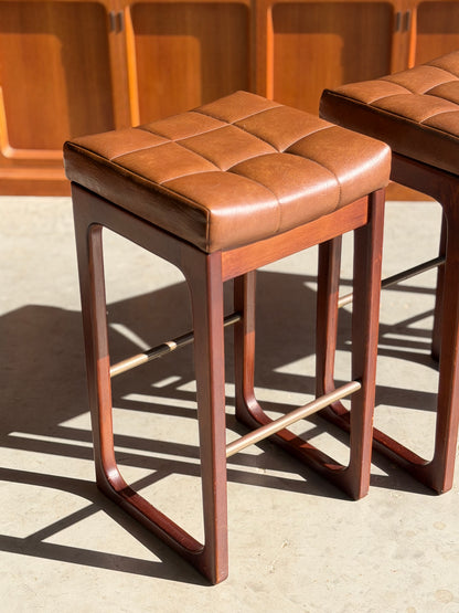 A set of three Mid Century Australian 1960s Barstools by Gerald Easden (Bar Height, Revive) with brown tufted leather seats, wooden legs, and metal footrests is shown on a sunlit concrete floor with wood paneling in the background.