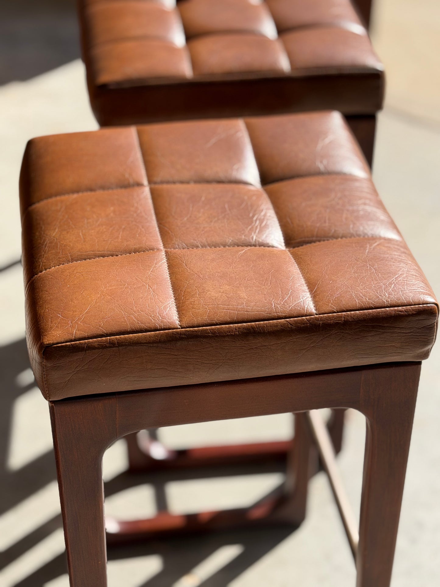A close-up of a brown, tufted leather Mid Century Australian 1960s Barstool by Gerald Easden (Bar Height, Revive) with a wooden base and sunlight across the seat; another stool from the set of 3 is slightly blurred in the background.