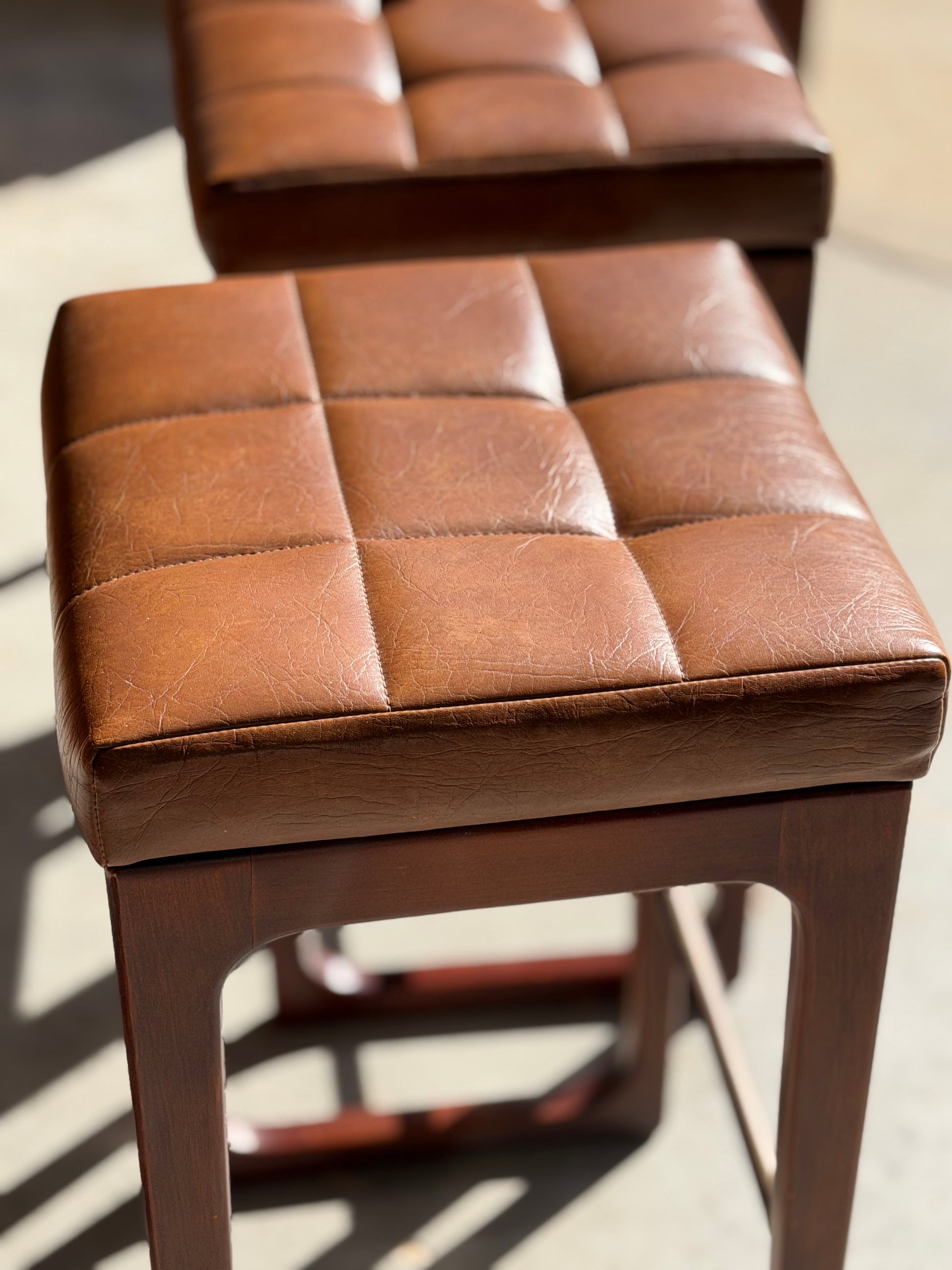 A close-up of a brown, tufted leather Mid Century Australian 1960s Barstool by Gerald Easden (Bar Height, Revive) with a wooden base and sunlight across the seat; another stool from the set of 3 is slightly blurred in the background.