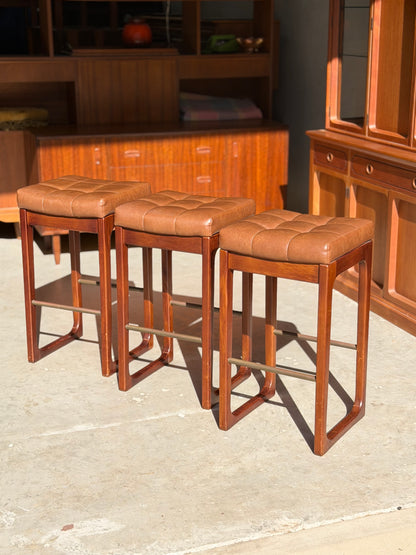 Three Mid Century Australian 1960s Barstools by Gerald Easden (Bar Height, Set of 3) are arranged in a row on a sunlit concrete floor, complemented by matching mid-century furniture and cabinets in the background.