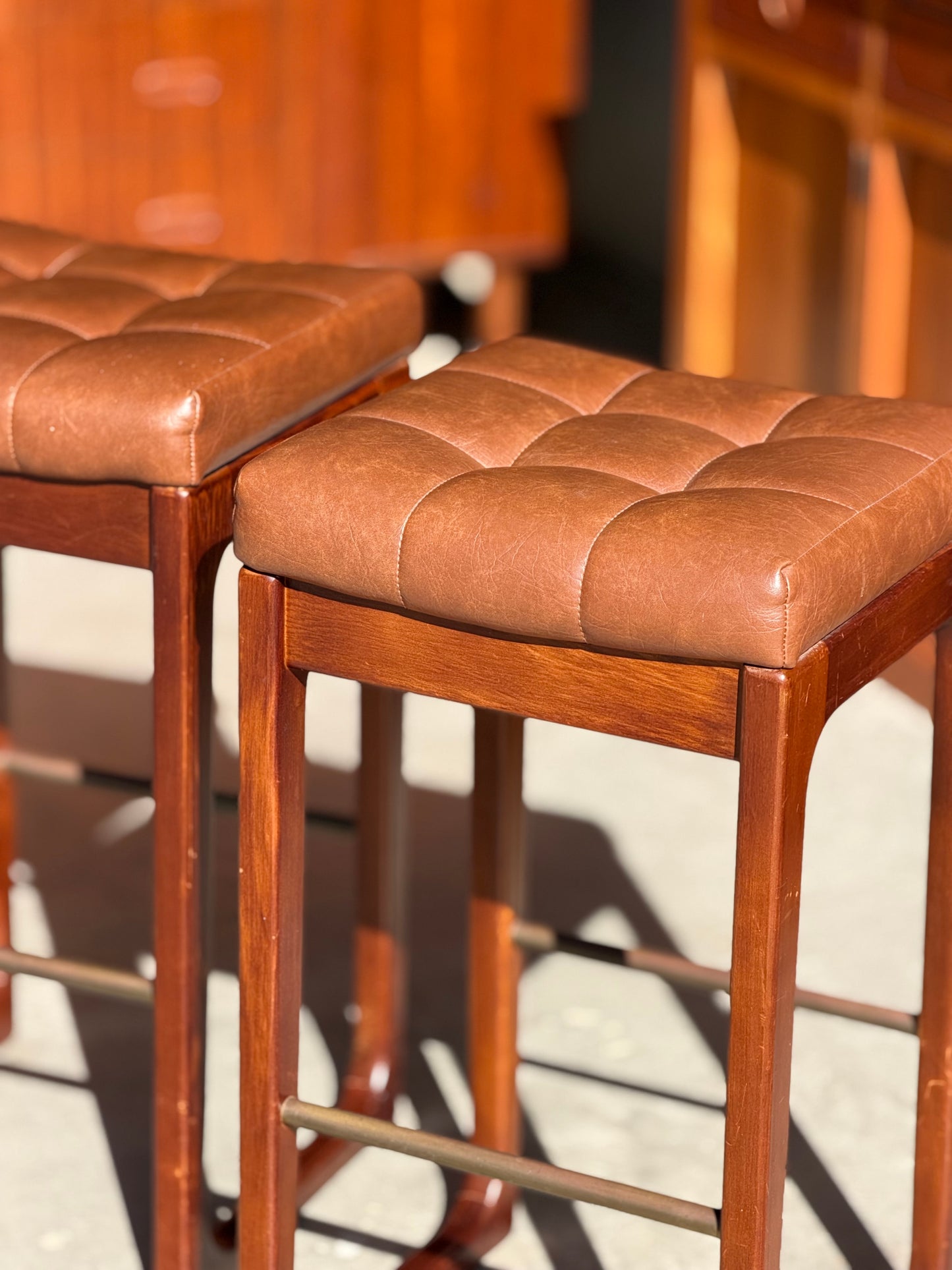 Three Mid Century Australian 1960s Barstools by Gerald Easden, bar height variant, feature square seats with brown tufted leather and sturdy wooden frames, capturing classic style with warm wood accents in bright sunlight.